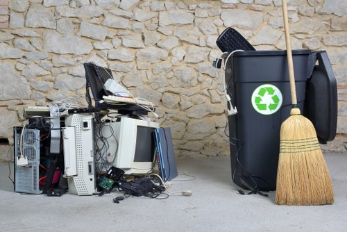 A pile of discarded electronic waste, including old computer towers, monitors, keyboards, and various cables, situated on a concrete surface in front of a textured stone wall. The items are stacked haphazardly with some cables hanging loose and parts leaning against each other, indicating they are intended for disposal or recycling. To the right, a large black rubbish bin with a green recycling symbol on its side is positioned on the ground, alongside a traditional straw broom with a wooden handle leaning against it. The scene suggests a typical waste collection or rubbish removal setting, likely in a residential or commercial area within Wandsworth, where Business Waste Removal Wandsworth provides services. The lighting appears natural, highlighting the textures of the stone wall and the various materials of the waste items, emphasizing the importance of proper rubbish removal and recycling practices in the local area.