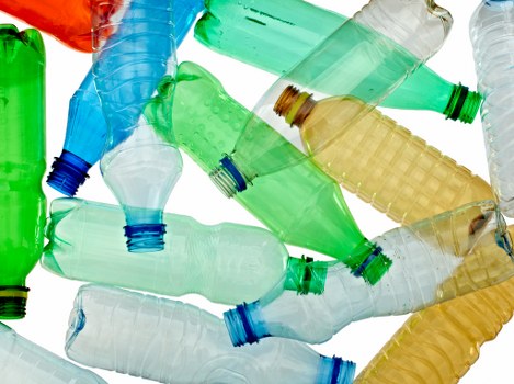 A collection of discarded plastic bottles in various colours including green, blue, and transparent, scattered across a surface. The bottles are made of PET plastic, with some showing ridged textures around the necks and bodies, and others with smooth finishes. Many bottles are crumpled, some with their caps removed or loosely attached, and they are positioned at different angles, overlapping each other. The background appears to be a clean, flat surface, possibly pavement or a work surface, with no other objects visible. The lighting is neutral, highlighting the transparency and muted colours of the bottles. The scene suggests an environment related to waste collection or recycling, aligning with the services of Business Waste Removal Wandsworth specializing in rubbish clearance services in the Wandsworth area.