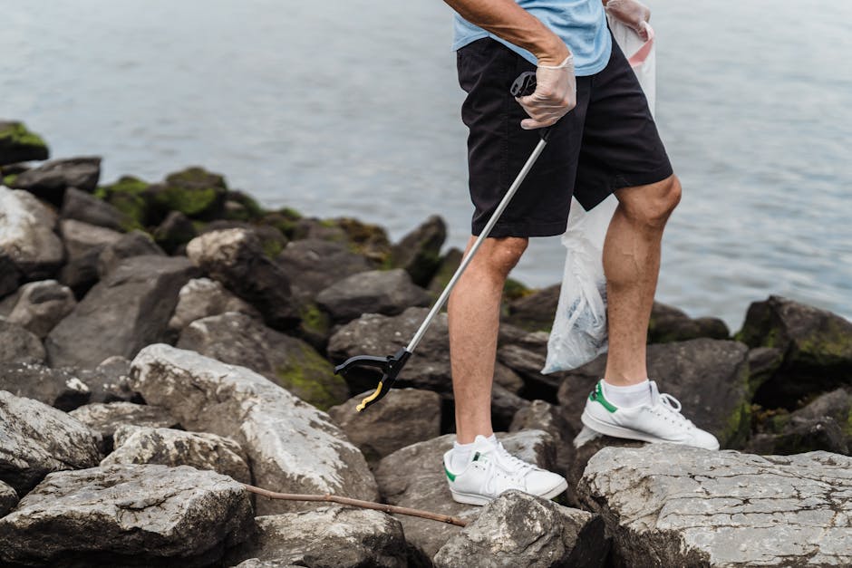 A person wearing grey gloves, a light blue T-shirt, black shorts, and white sneakers with green accents is standing on a rocky shoreline near water, holding a trash picker tool in their right hand and