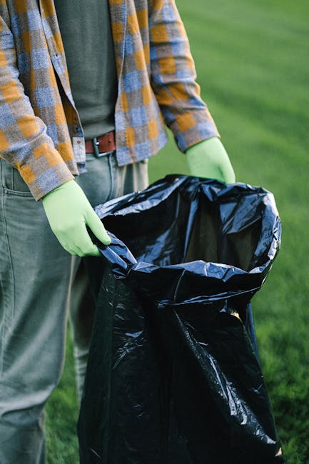 A person wearing grey gloves, a light blue T-shirt, black shorts, and white sneakers with green accents is standing on a rocky shoreline near water, holding a trash picker tool in their right hand and