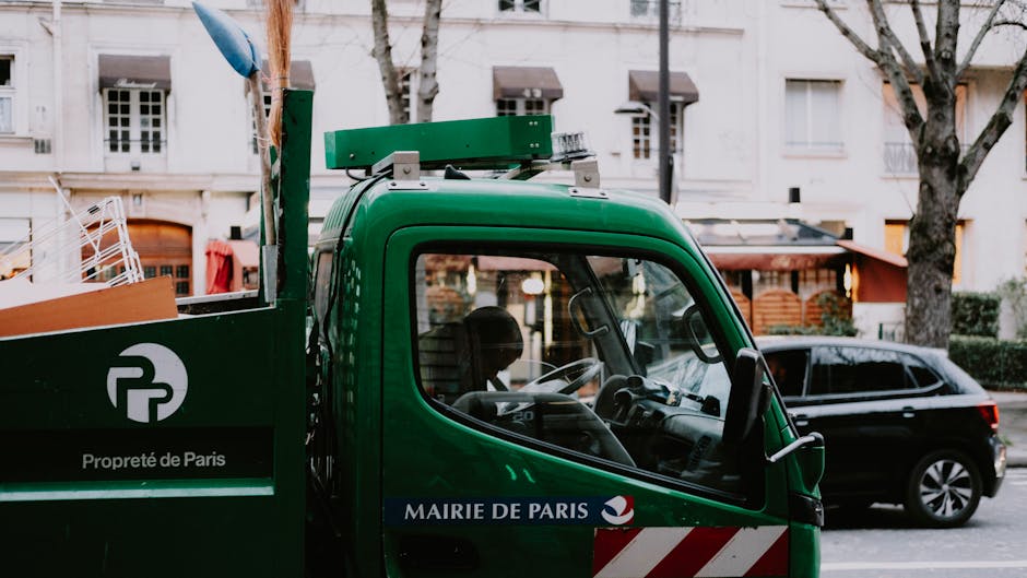 A close-up view of a green waste collection truck parked outdoors, with the driver's side window visible. The truck has a black and white logo with the text 'Propreté de Paris' on a green panel, and a