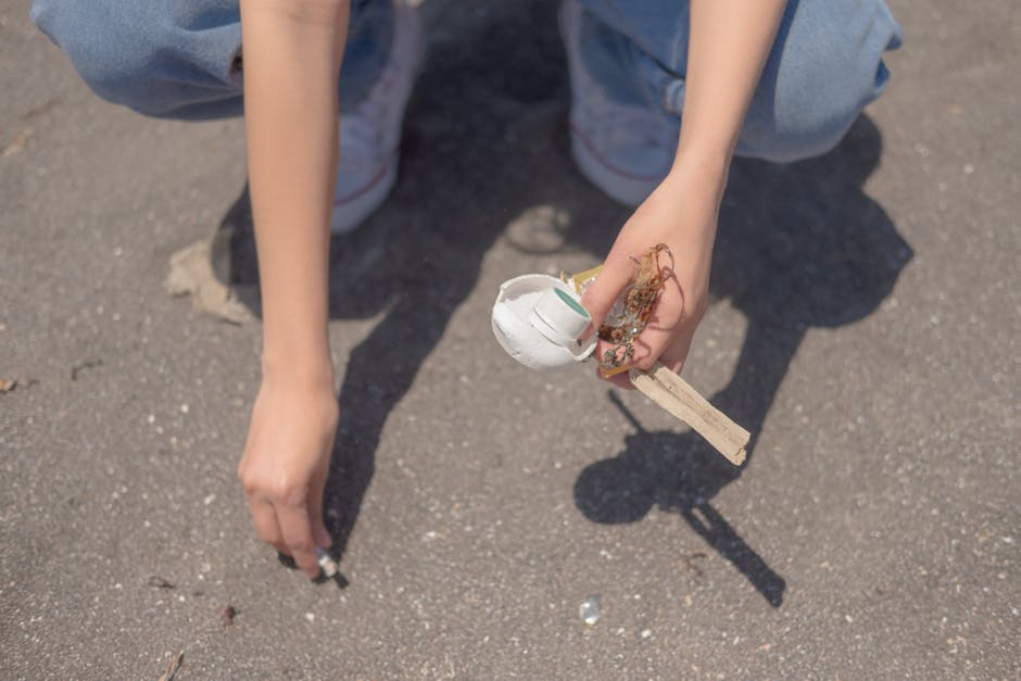 A person dressed in blue jeans and white sneakers crouches on a paved outdoor surface, holding a small collection of discarded waste. Their right hand is gripping a makeshift construction of a wooden