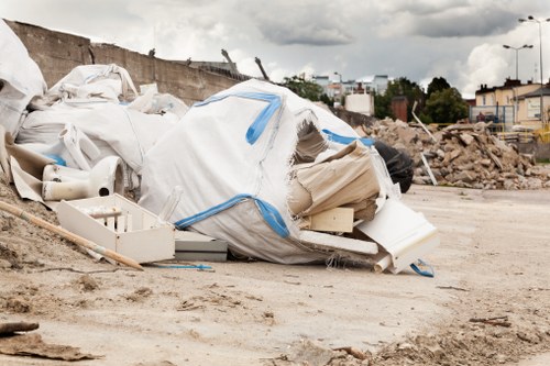 A large pile of mixed waste materials including white plastic bags, some torn open revealing containers and packaging, is situated on a dirt and gravel surface in an outdoor area. The waste appears provisional and is piled loosely with some items spilling onto the ground. In the background, there is a partially demolished concrete structure, construction debris, and a busy urban setting with buildings, trees, and streetlights visible under a cloudy sky. The scene suggests a site undergoing clearance, where waste removal services such as those provided by Business Waste Removal Wandsworth operate to manage rubbish collection and disposal in the Wandsworth area of London. The overall environment indicates an active construction or renovation site preparation, emphasizing the need for efficient rubbish management.