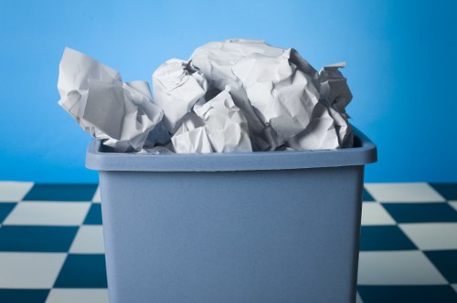 A light blue plastic bin filled with tightly crumpled sheets of white paper, placed on a black and white checkered tiled floor with a solid light blue background. The bin appears to be used for waste disposal or rubbish collection, with the crumpled paper occupying the majority of the container's opening. The setting suggests an indoor environment, possibly a utility or storage area, associated with rubbish removal services in Wandsworth, as indicated by the website domain. The paper's texture is uneven with visible creases and folds, and the bin's surface is smooth with a matte finish. This image illustrates typical rubbish container use, relevant to professional waste management and rubbish removal services offered by Business Waste Removal Wandsworth, focusing on the collection of paper waste.